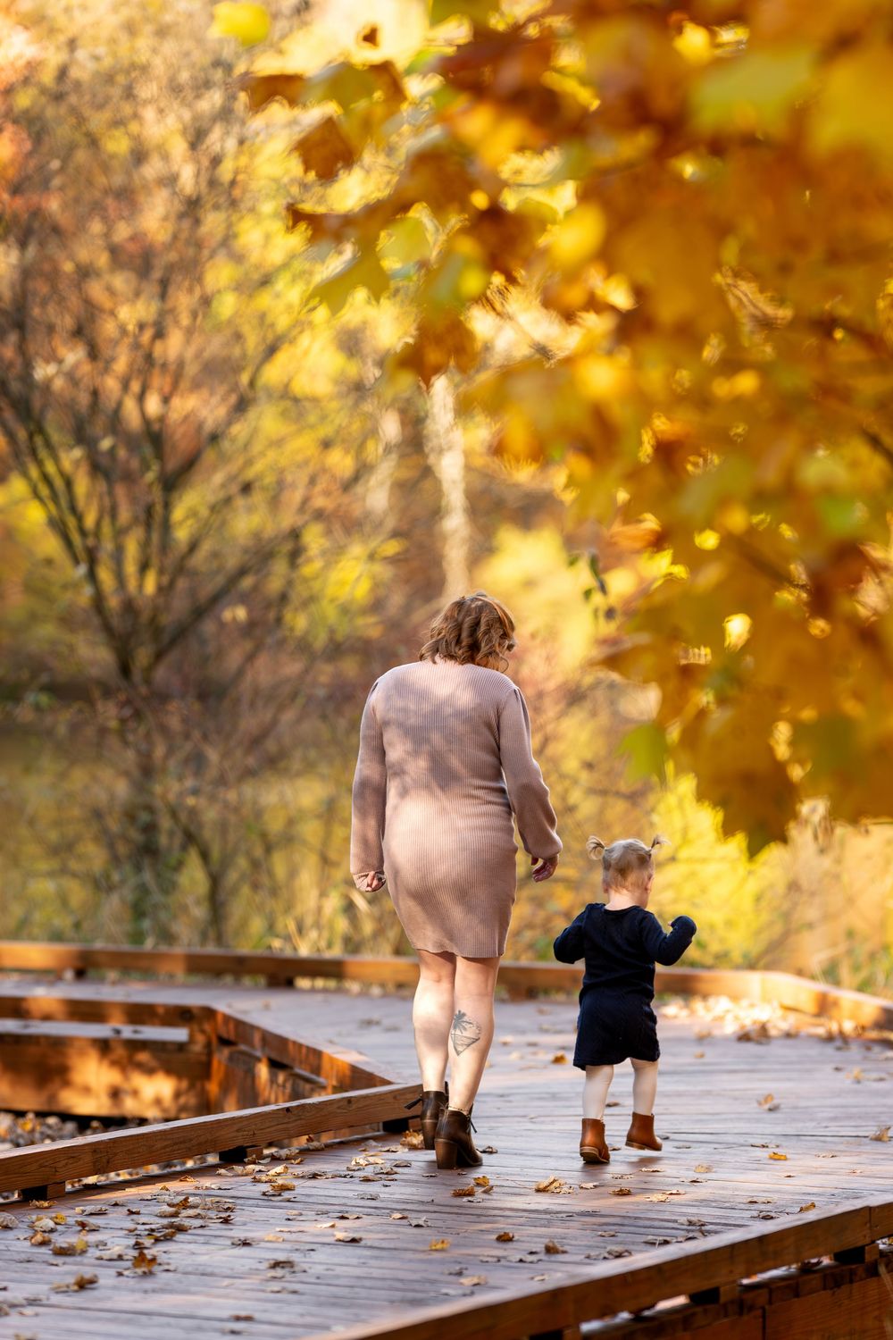 Pregnant mother and toddler walking hand-in-hand on wooden bridge in fall forest.