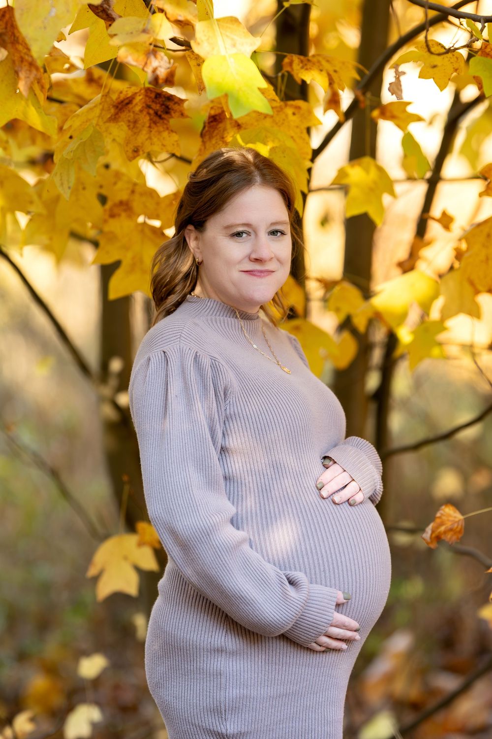 Smiling pregnant woman in taupe sweater dress cradling baby bump surrounded by golden fall leaves.