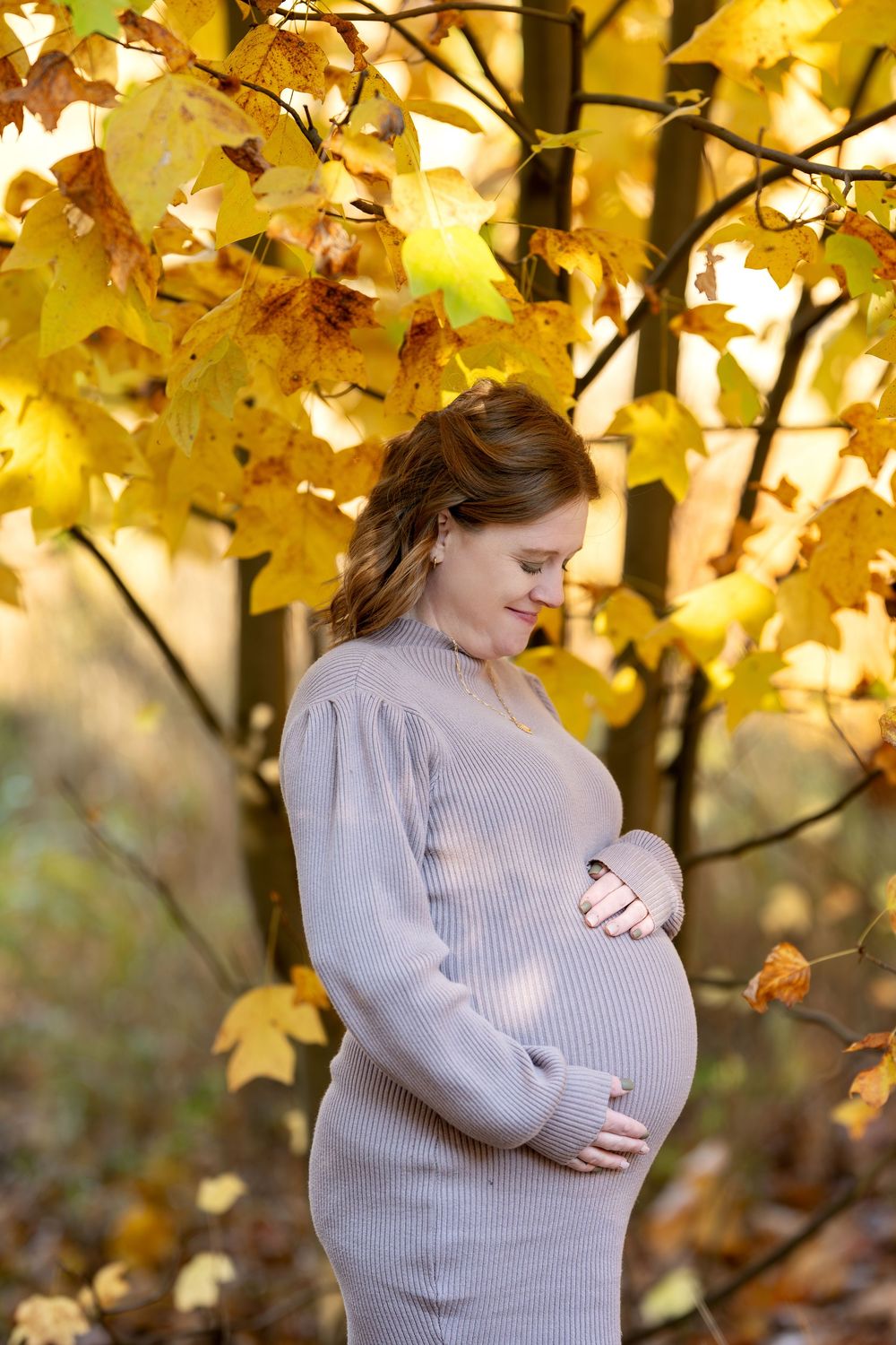 Pregnant woman in beige knit dress cradling belly under golden autumn maple tree.