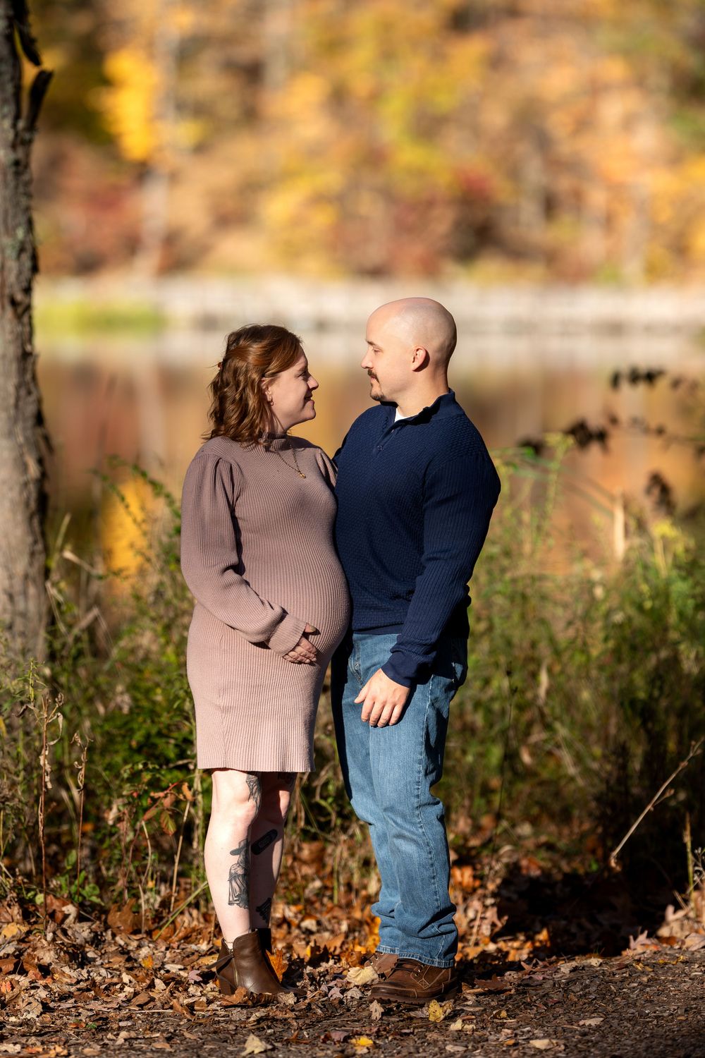 Expecting couple sharing a tender moment by a lake surrounded by fall foliage.