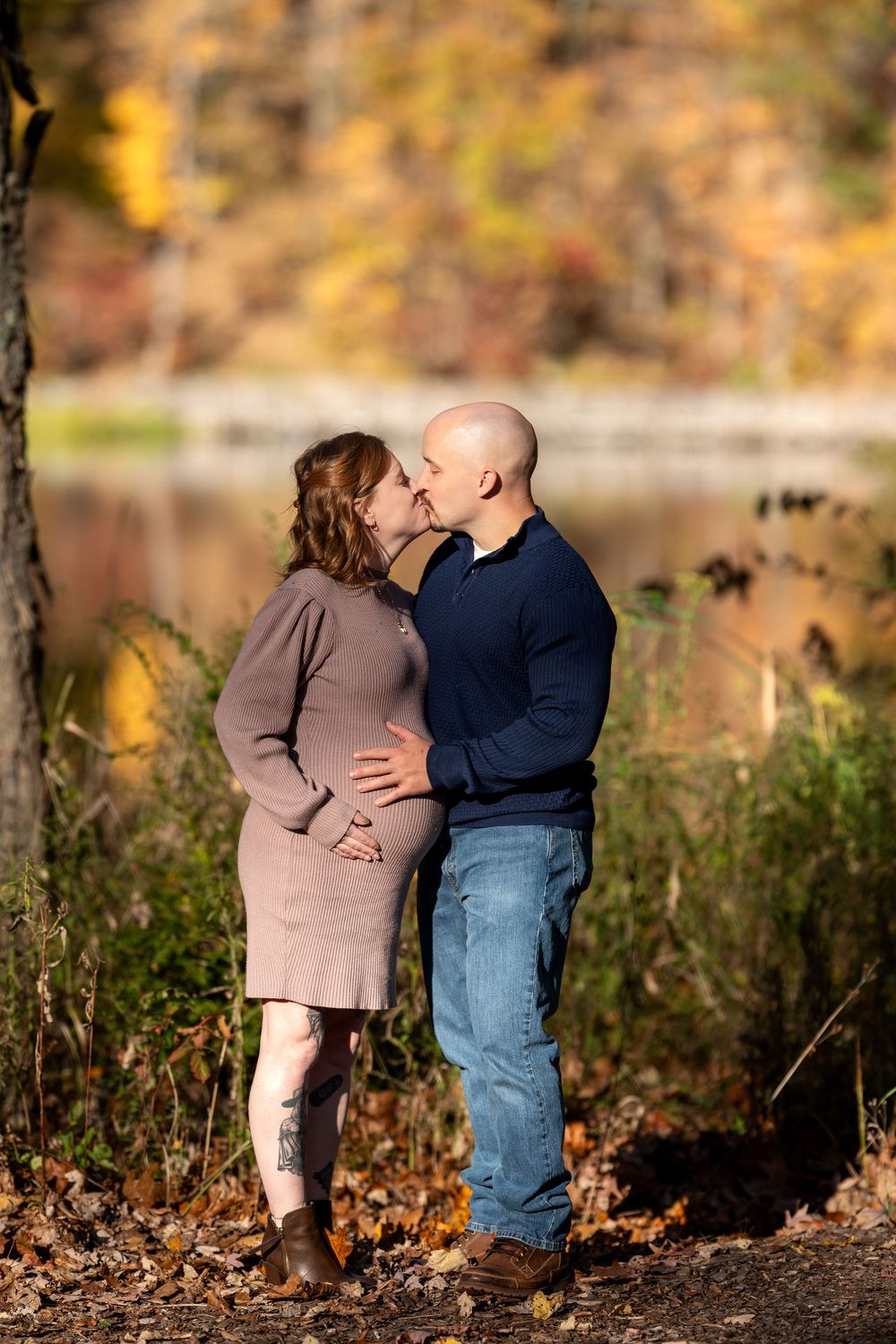Pregnant couple sharing a kiss outdoors amid vibrant autumn colors near a lake.