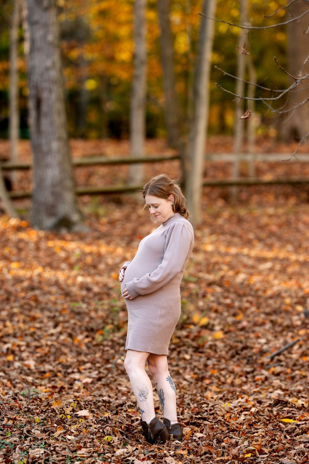 Expectant mother in taupe dress looking down at baby bump surrounded by golden fall foliage.