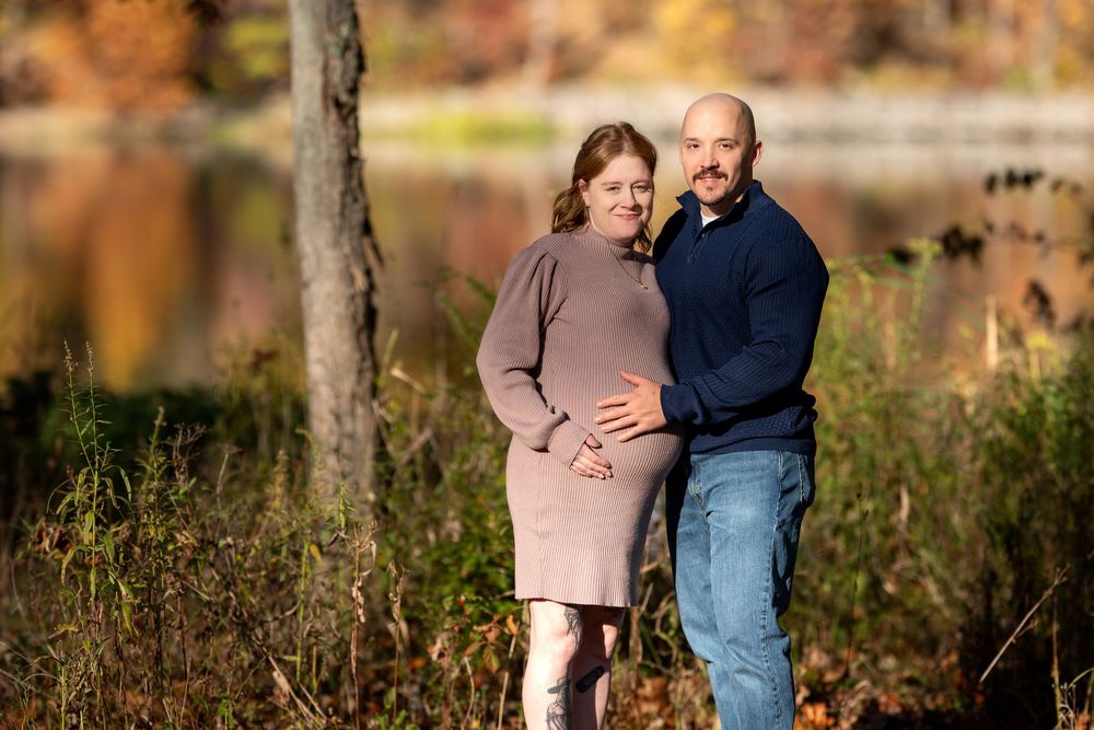 Pregnant woman and partner embracing outdoors in a colorful autumn forest by a lake.