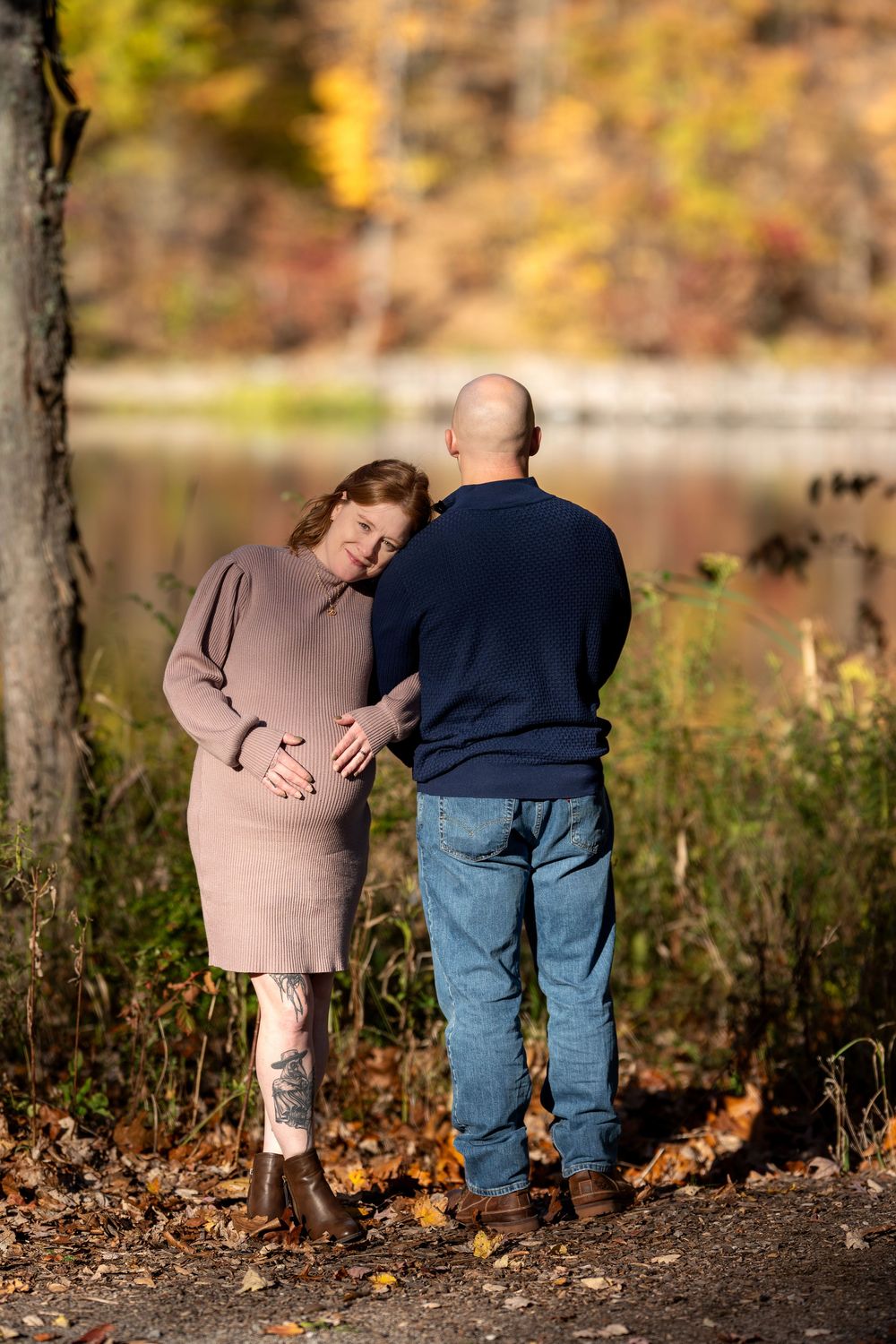Expectant couple standing back-to-back near a serene lake surrounded by fall foliage.
