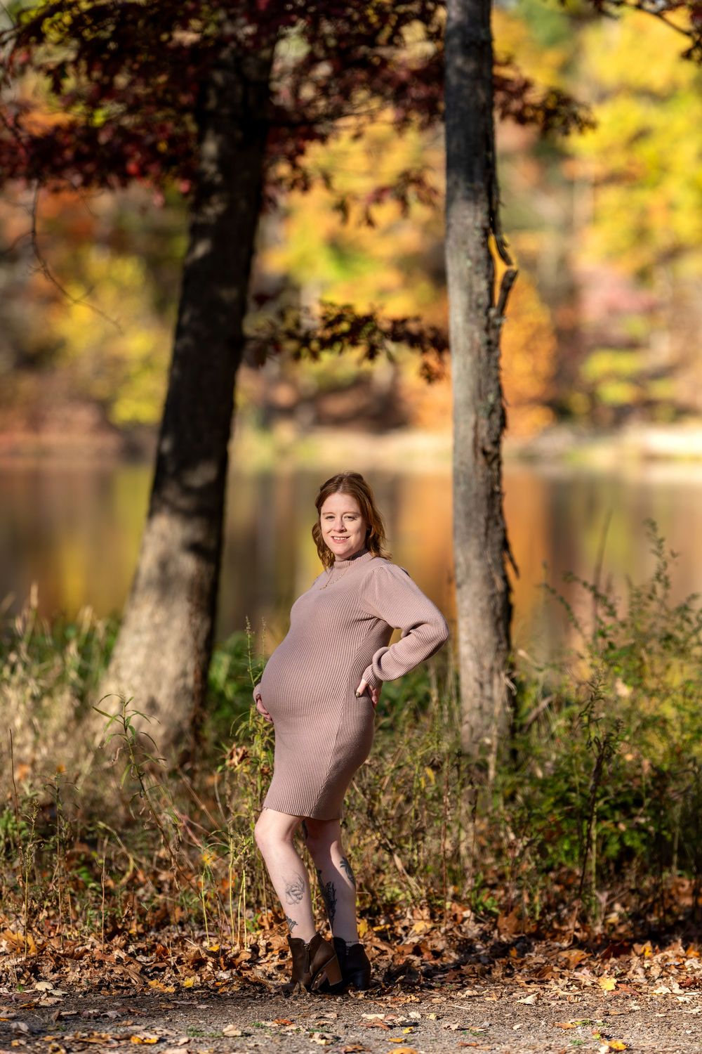 Pregnant woman in beige dress posing between trees with autumn lake backdrop.