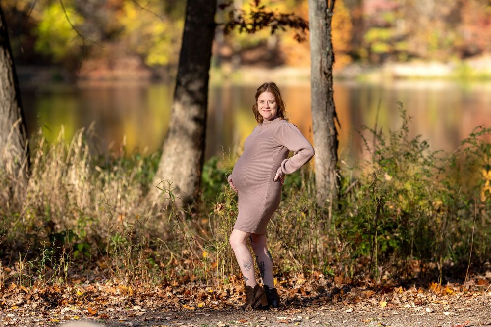 Smiling pregnant woman in fitted beige dress standing in sunlit autumn forest by lake.