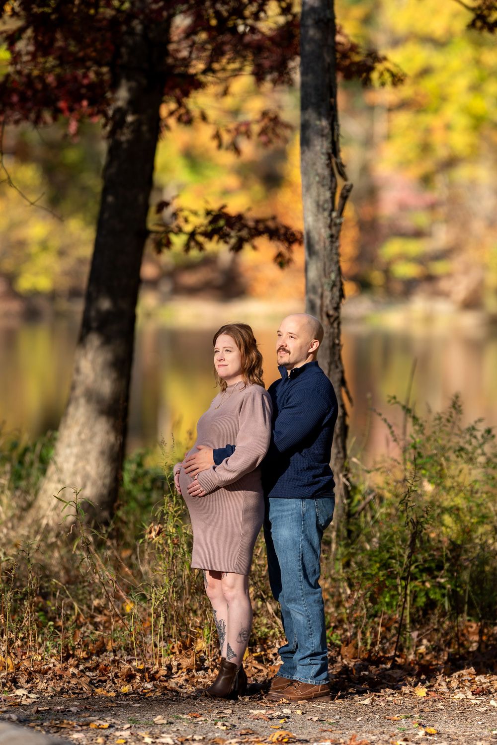 Expecting couple standing back-to-back by a serene lake surrounded by vibrant fall foliage.
