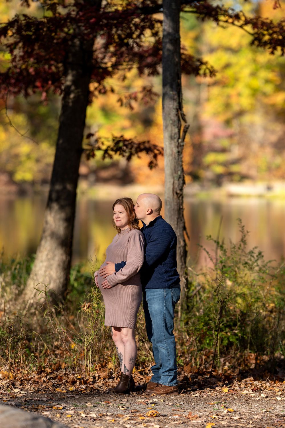 Couple sharing tender moment in autumn park, man kissing pregnant woman's cheek near colorful fall foliage and lake.