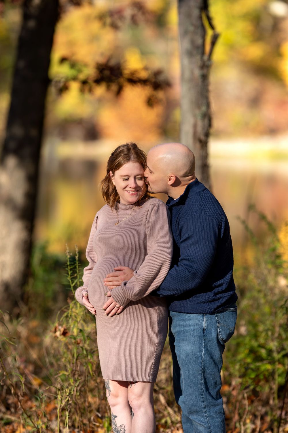 Man kissing smiling pregnant woman's cheek in vibrant fall park setting with golden foliage background.