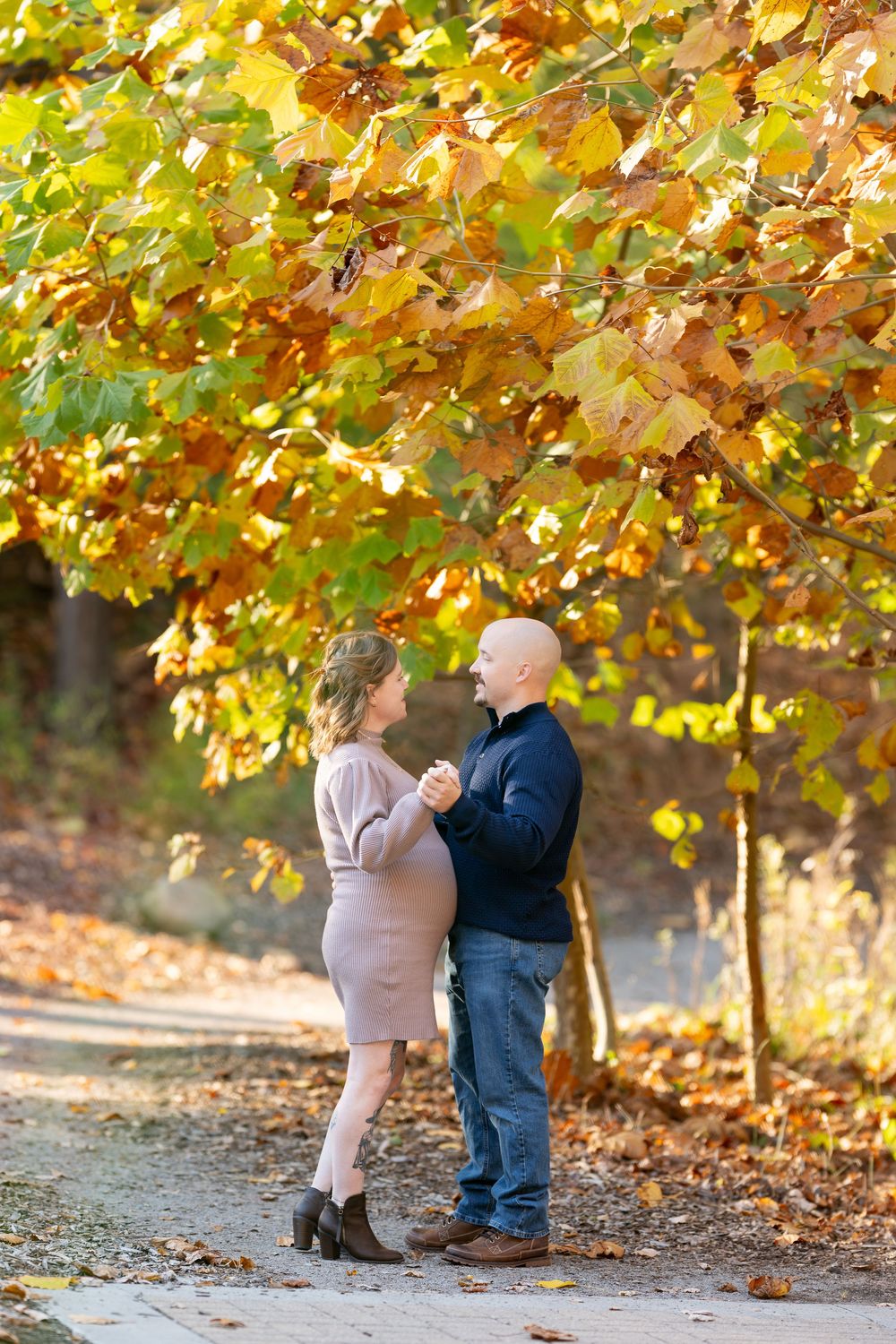 Pregnant couple holding hands and dancing on path under brilliant yellow autumn tree canopy.