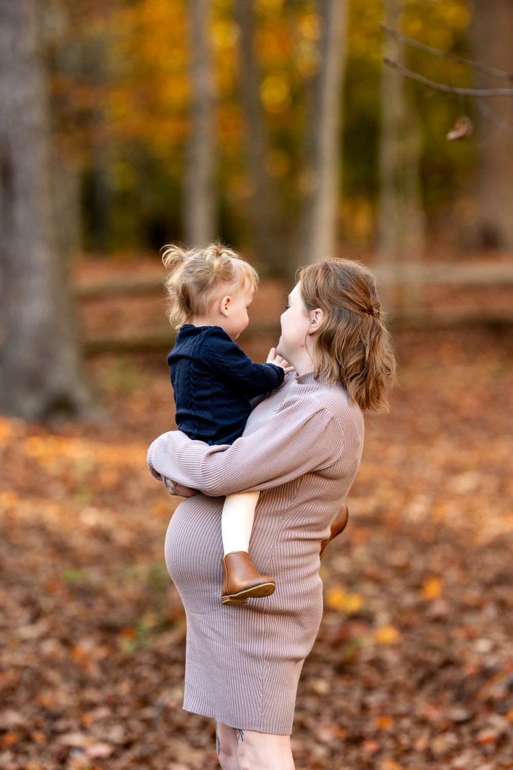 Pregnant woman in taupe dress holding toddler girl face-to-face in autumn forest setting.