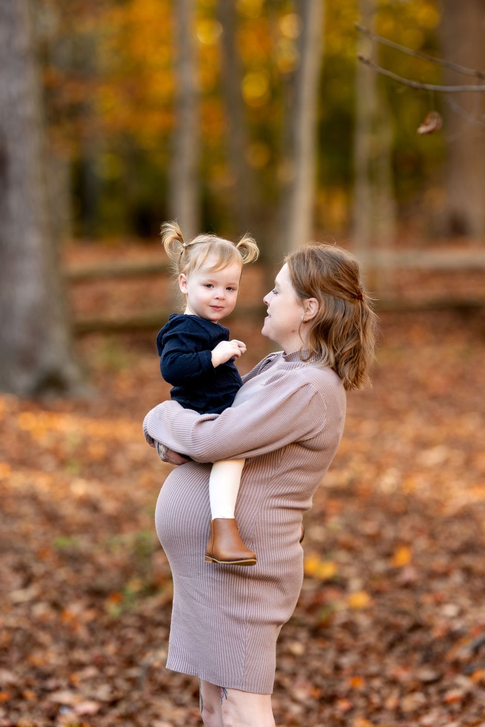 Smiling pregnant mother holding young toddler daughter in fall woodland with golden leaves.