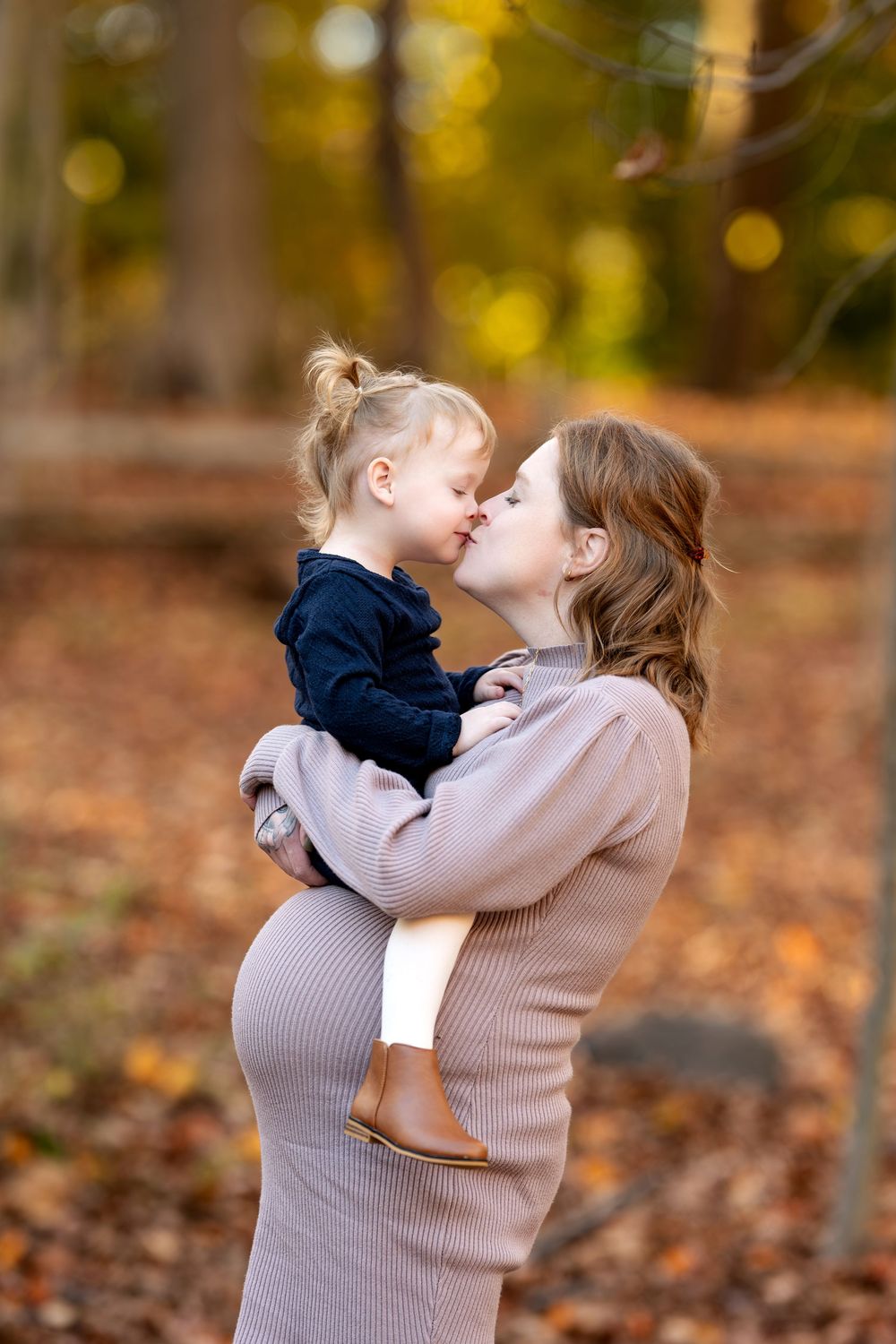 Pregnant woman in beige dress kissing toddler girl in autumn forest.