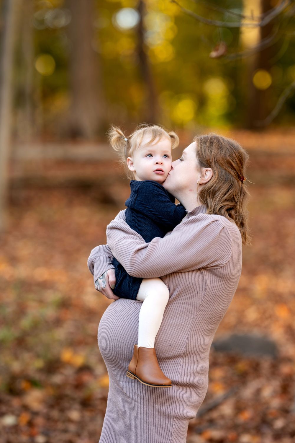 Pregnant mother kissing toddler daughter on cheek in fall woodland setting.