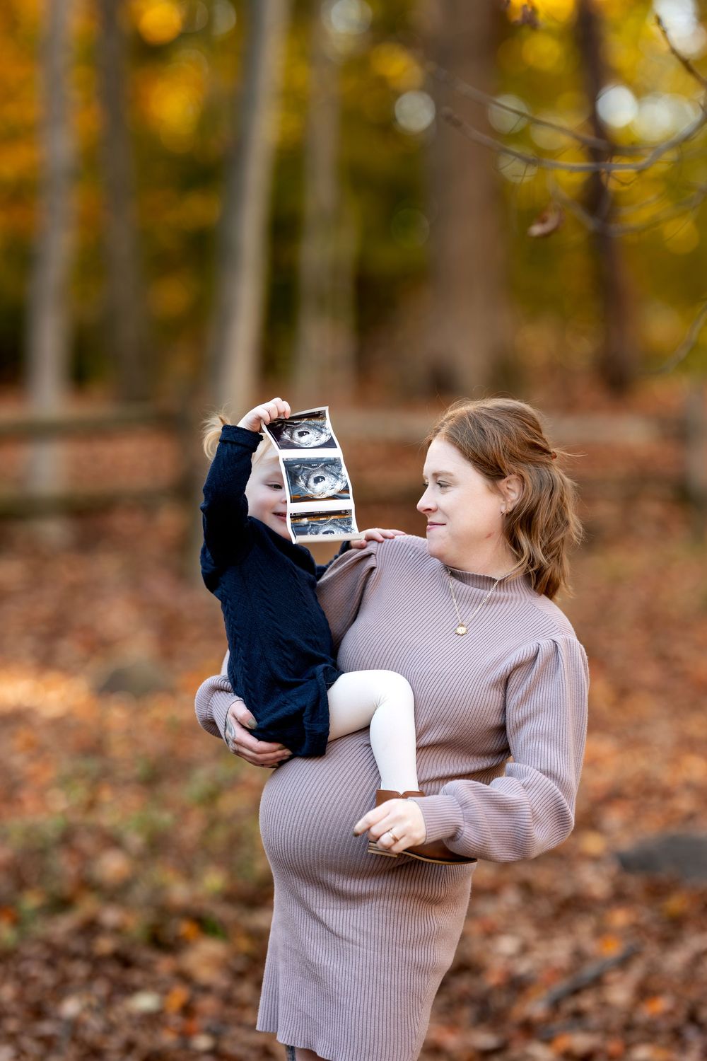 Pregnant woman holding child displaying ultrasound images in colorful fall forest.