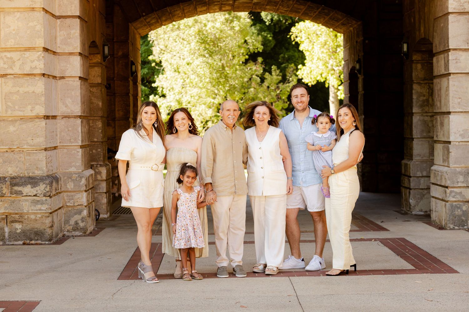 A group in coordinated light-colored outfits poses together under a stone archway on a sunny day.