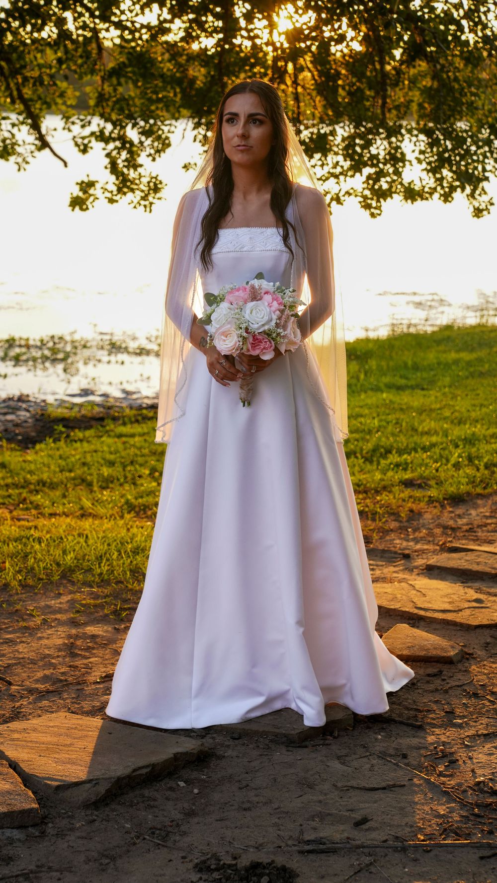 Bride in white satin gown holding pink bouquet standing outdoors near lake at golden hour sunset.