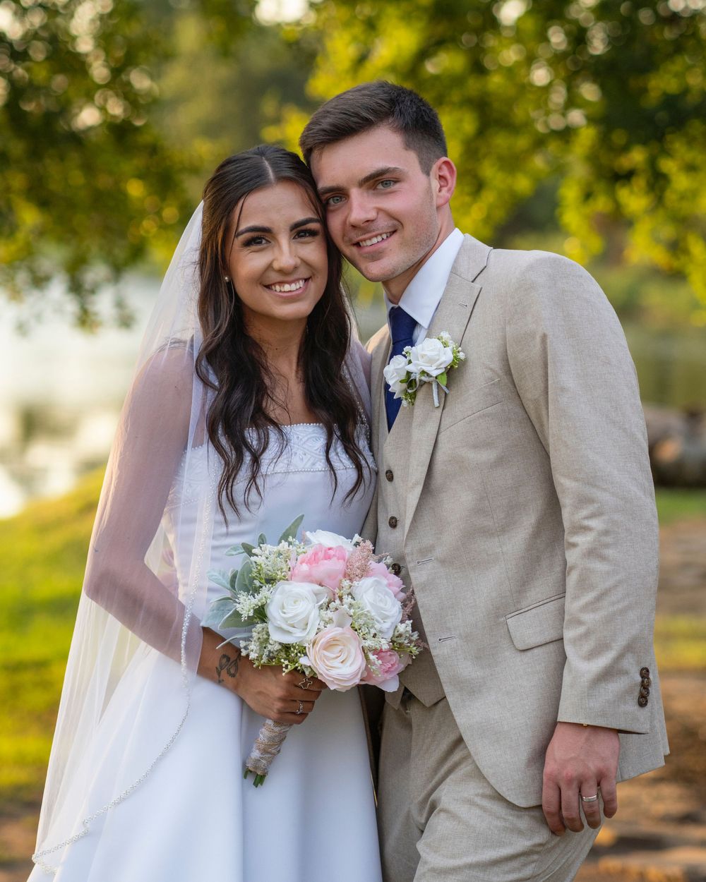 Smiling newlyweds pose outdoors, bride in white gown with veil holding pink bouquet, groom in beige suit.