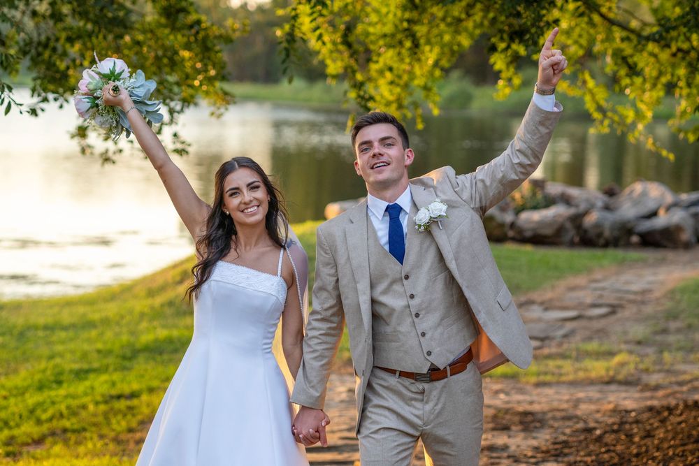 Happy newlyweds celebrating outdoors by lake at sunset, groom in tan suit, bride in white dress.