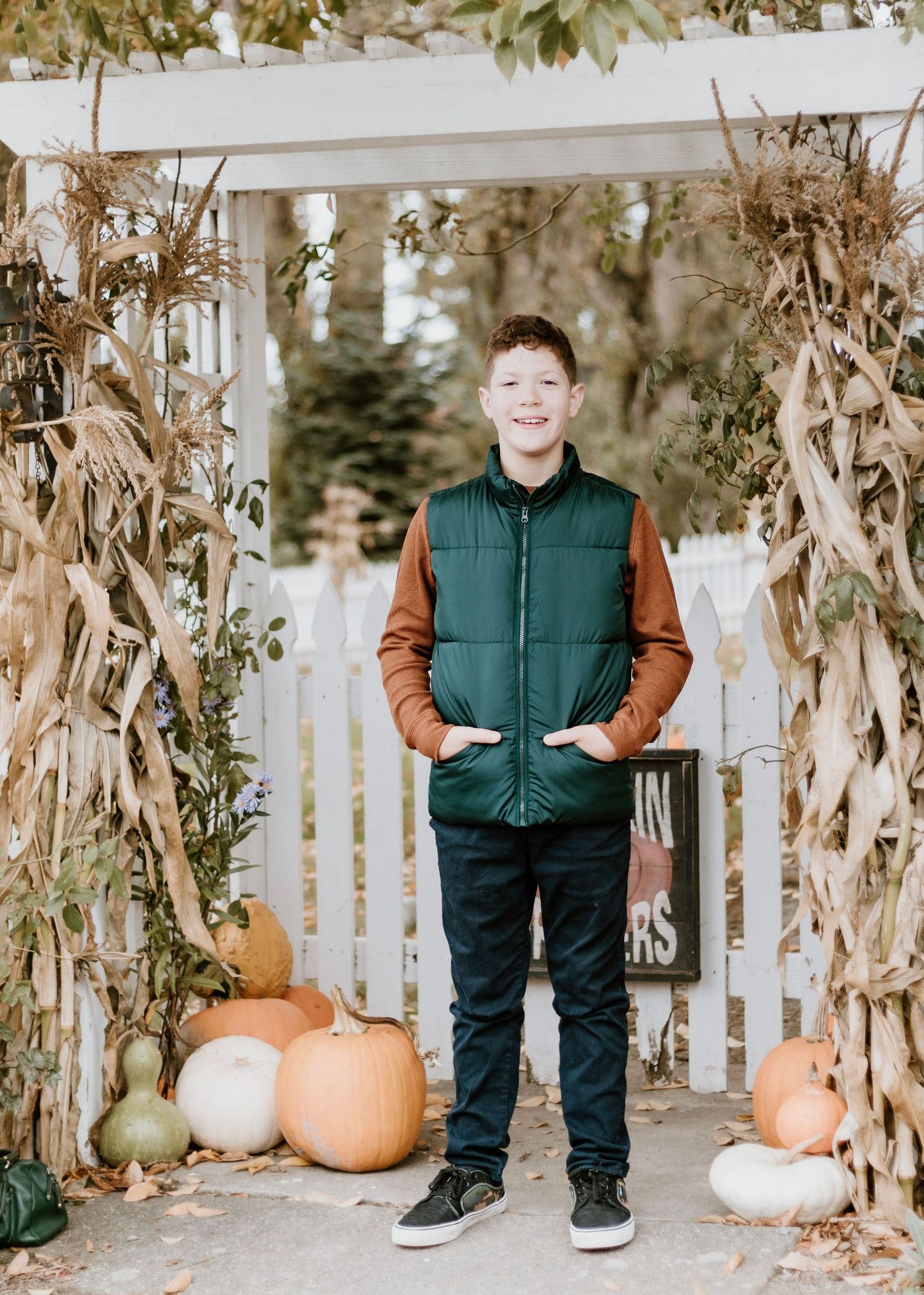 Fall portrait of young person in green vest standing among cornstalks and pumpkins.