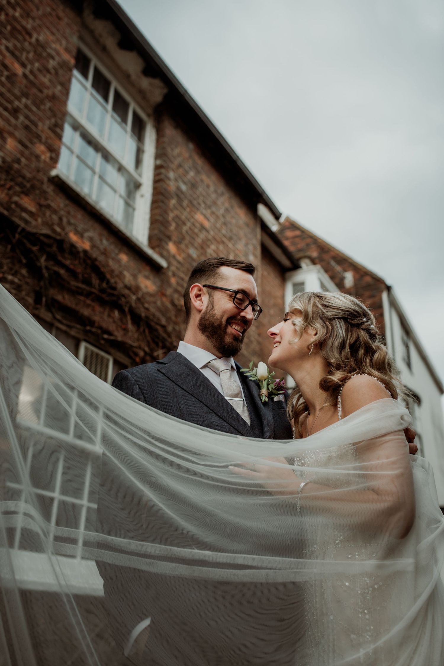 A joyful couple shares an intimate moment under a flowing veil against a rustic brick building backdrop.