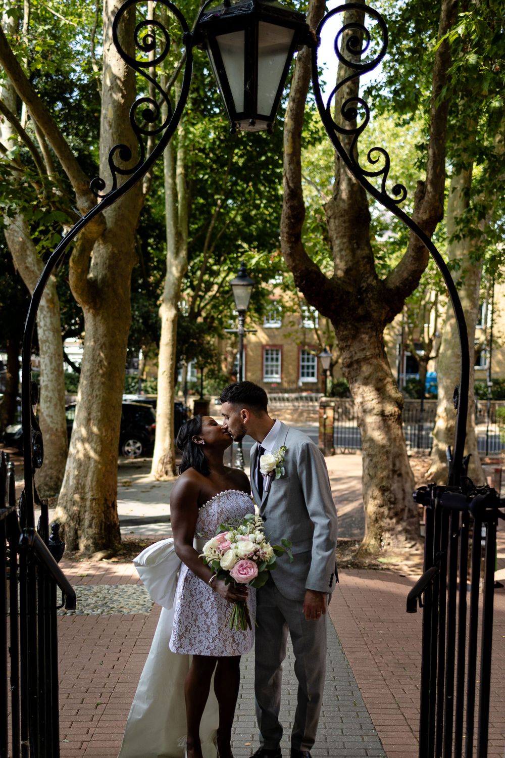 A newlywed couple shares a romantic moment under an ornate lamp post in a park setting.