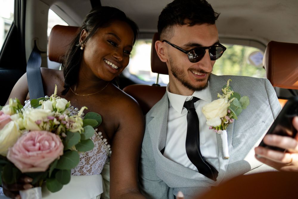 A newlywed couple looks at a phone together in the back of a car with pink roses in the bouquet.