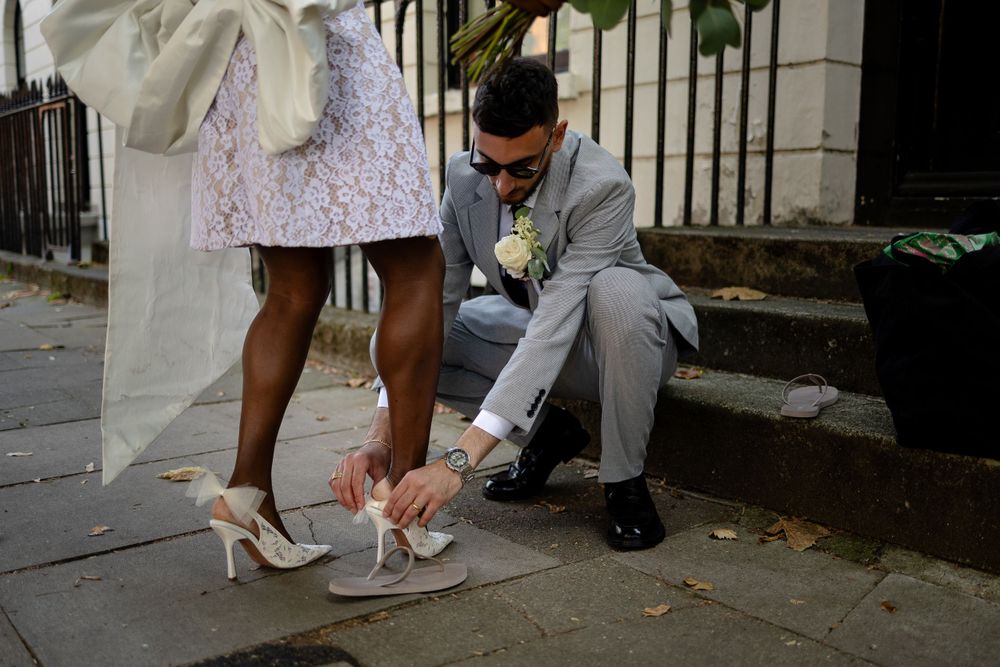 A groom helps adjust the bride's white high heel shoe on outdoor steps.