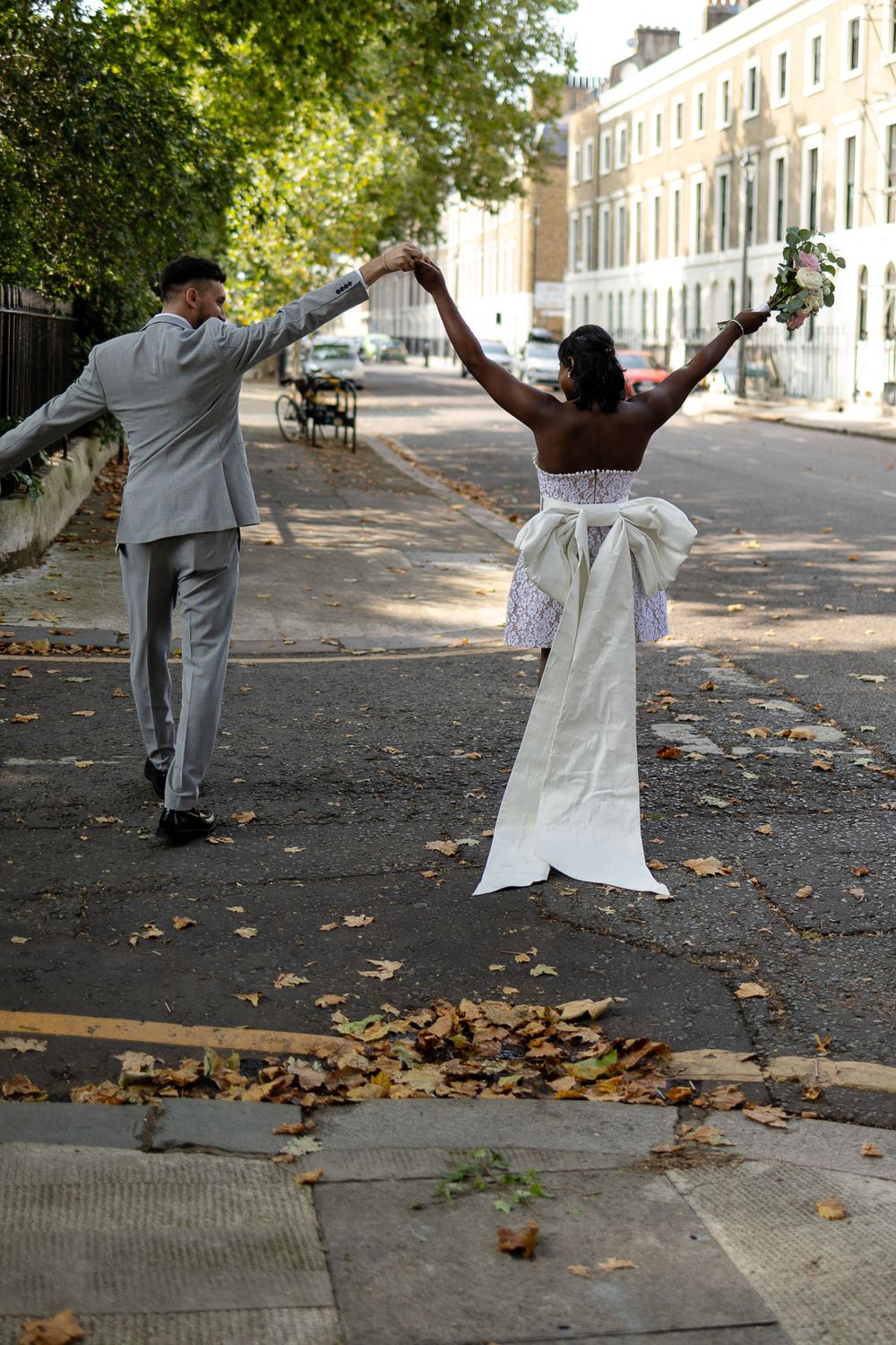A joyful couple dances and twirls on a tree-lined street in wedding attire.