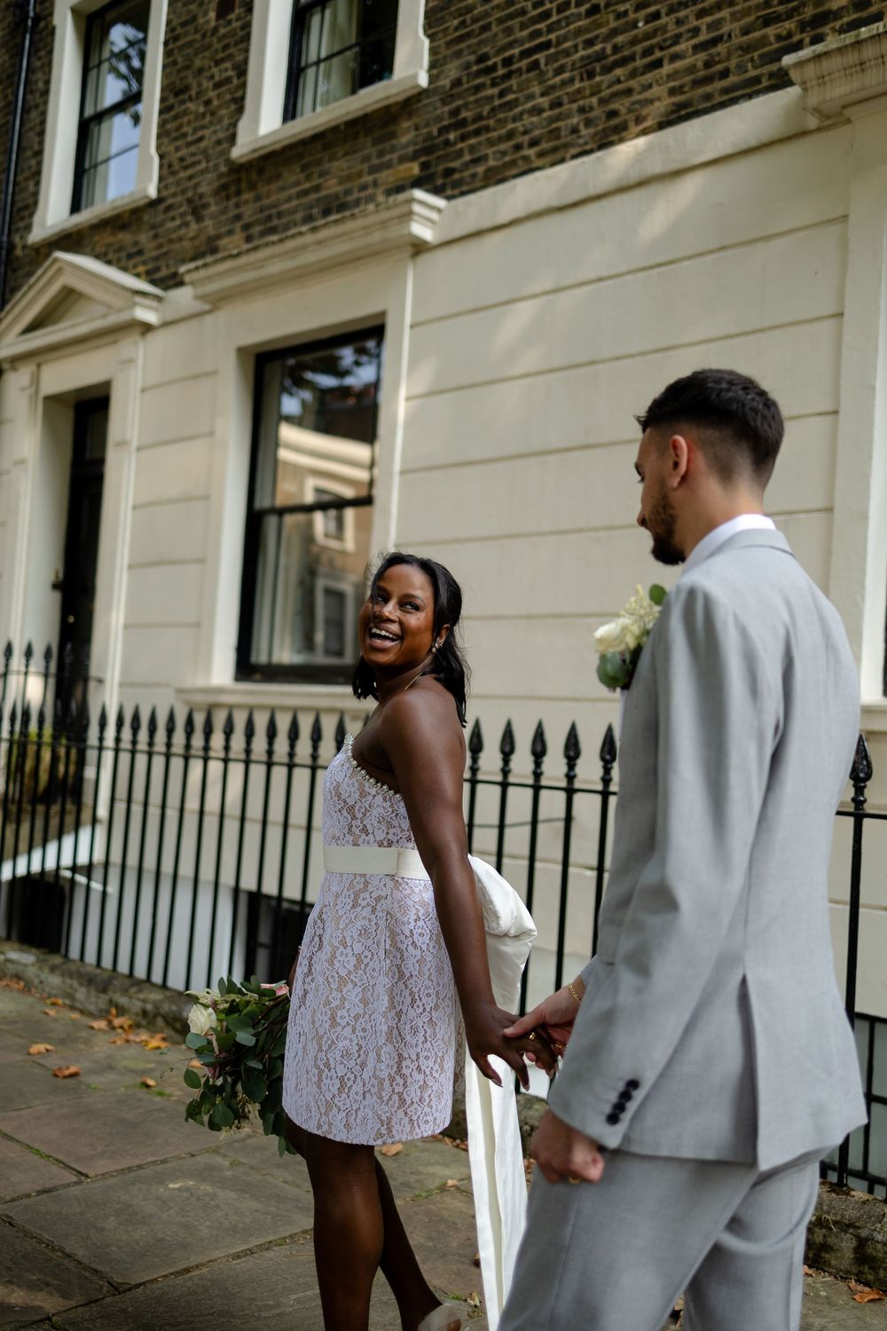 A bride in a lace dress looks back while standing by an elegant townhouse facade.