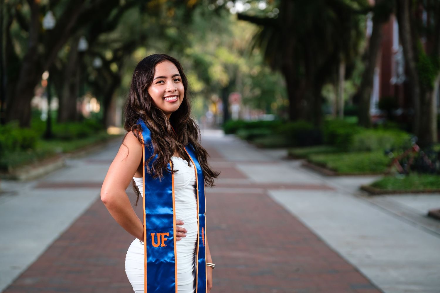 A smiling UF grad wearing a white dress and blue stole stands on a brick, tree lined walkway at the University of Florida.