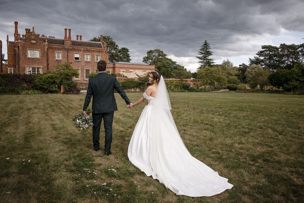 A couple in wedding attire walks across a scenic hilltop lawn on an overcast day at an elegant estate property.