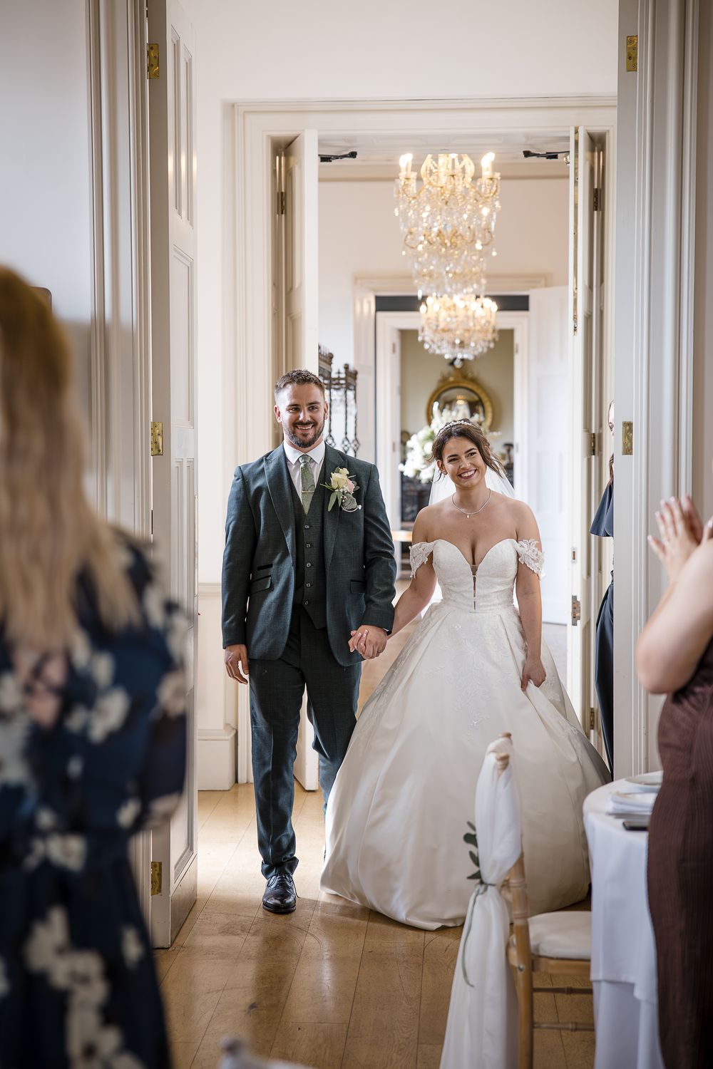 Wedding party walking through bright hallway with ornate chandeliers and white walls.