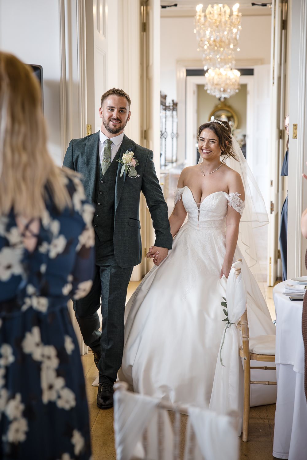 Wedding couple walking through an elegant ballroom with crystal chandeliers and white decor.