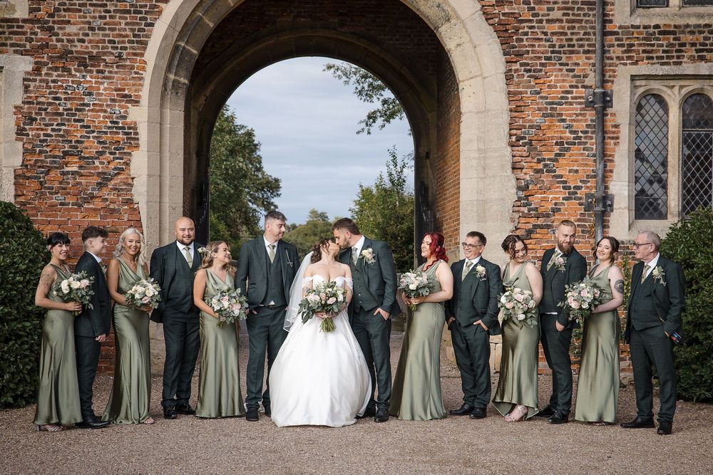 Wedding group photo under brick archway of historic building with bridesmaids in sage dresses.
