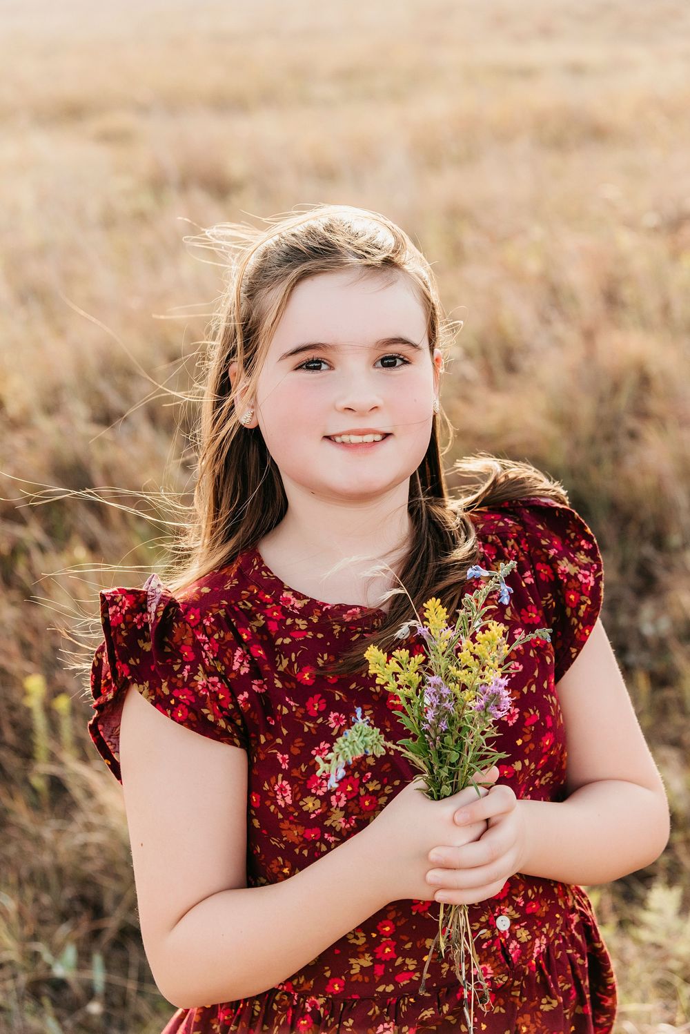 Young child in red velvet dress holds wildflowers while posing in sunlit meadow.