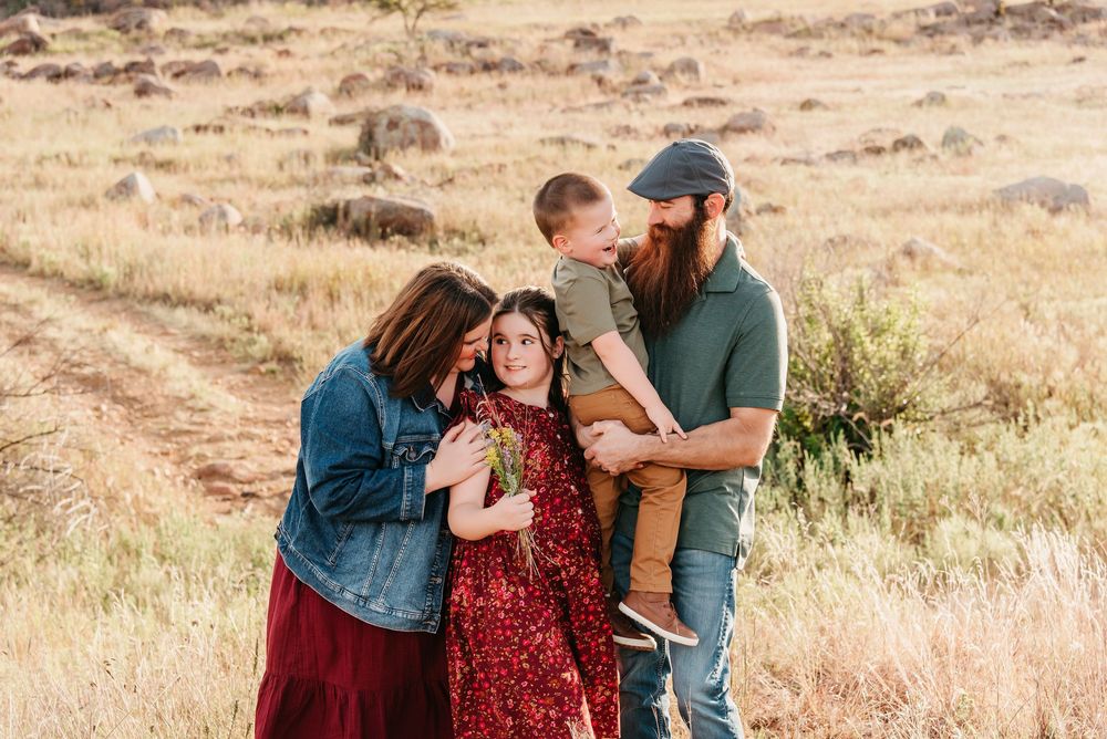 A couple embraces in a golden field during sunset while wearing burgundy and denim outfits for their engagement photos.