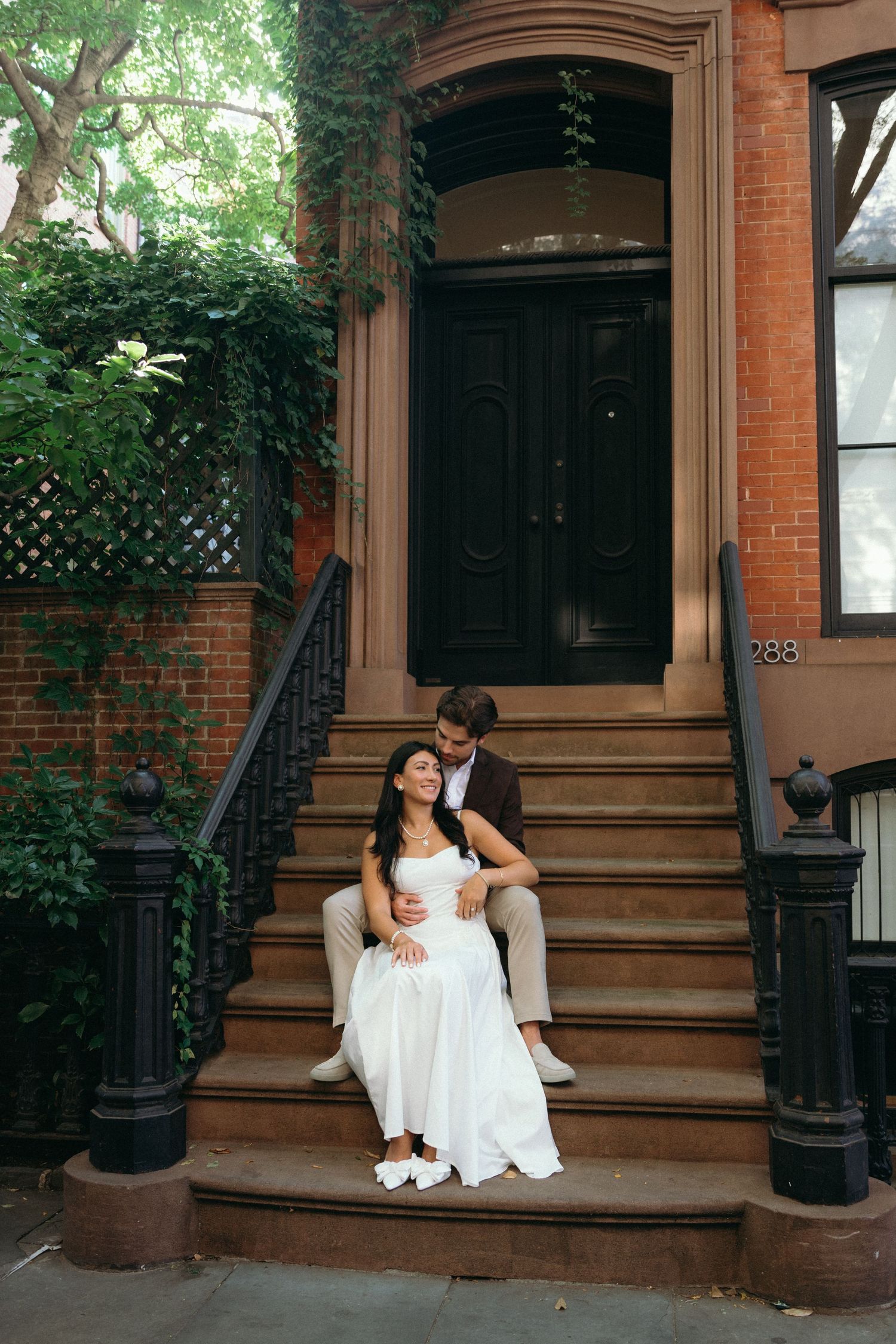 A couple sits together on the steps of a historic brownstone building surrounded by greenery and brick architecture.