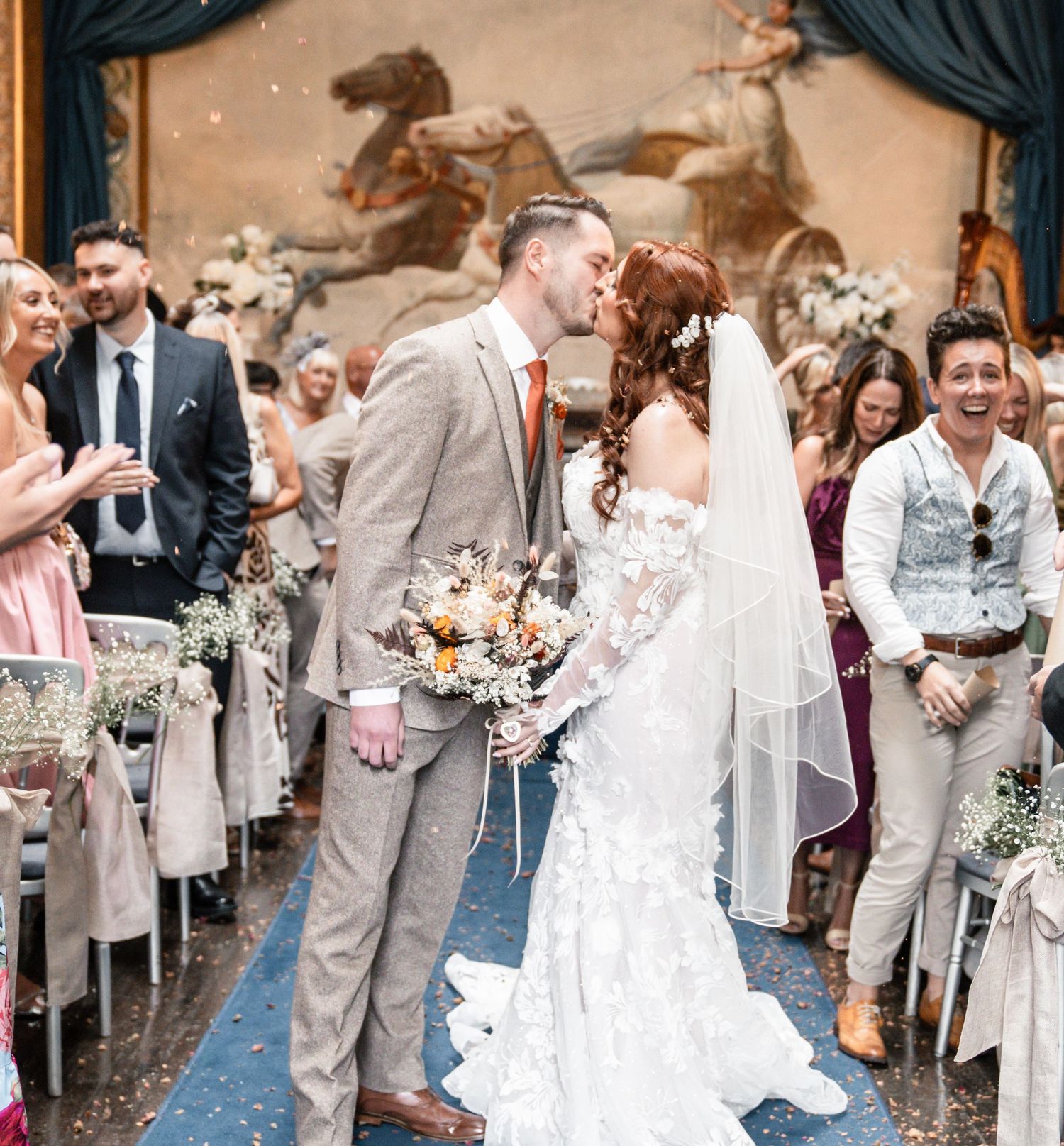 A joyful wedding celebration shows a couple sharing their first kiss as guests cheer during an indoor ceremony.