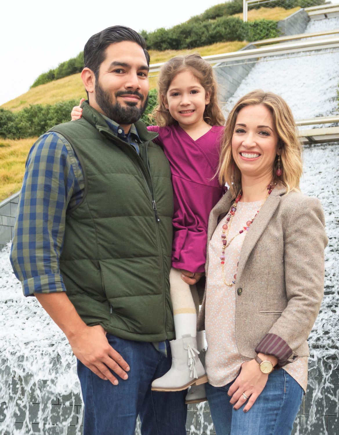 Family of three in front of a waterfall at a park, warm candid moment in natural light.