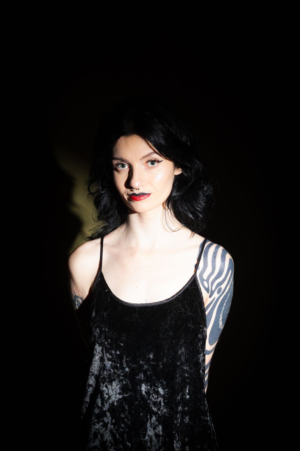 A figure in a black velvet tank top with dark hair and visible arm tattoo poses against a dark background.
