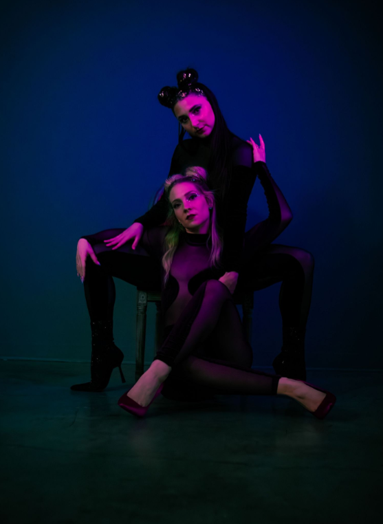 Two dancers in dark outfits strike a dramatic pose under purple and blue lighting on a dark stage.