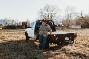 A cowboy in a plaid shirt and hat leans against a flatbed pickup truck in a rural field.