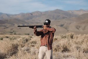 Person aiming a rifle in a desert landscape with mountains in the background.