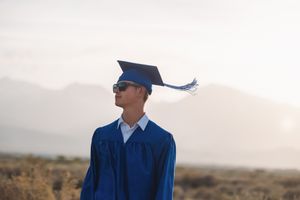 Young male graduate in blue cap and gown with sunglasses poses outdoors with mountains in background.