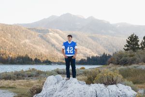 Teen boy in blue #42 football jersey stands on rock with mountain lake landscape backdrop.