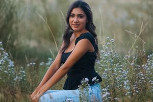 Woman in black tank top and jeans sitting among wildflowers in natural setting.
