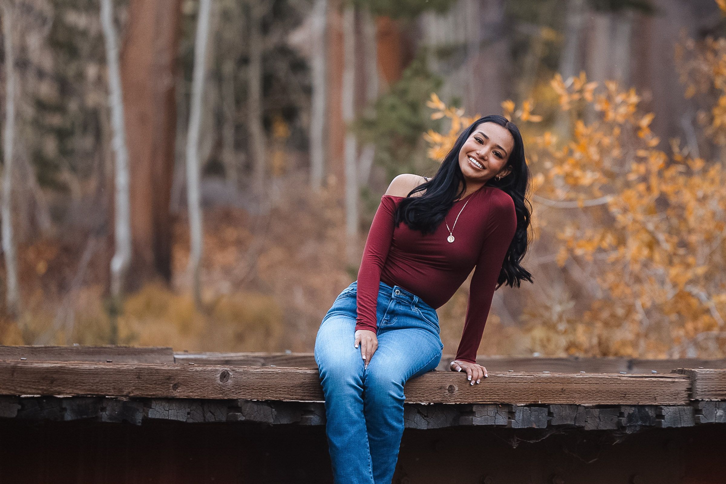 Person in casual attire posing playfully on wooden bench with autumn trees in background.