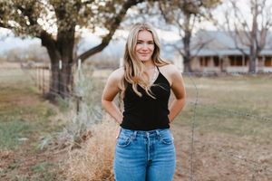 Woman in black top and blue jeans posing outdoors in rustic country setting.