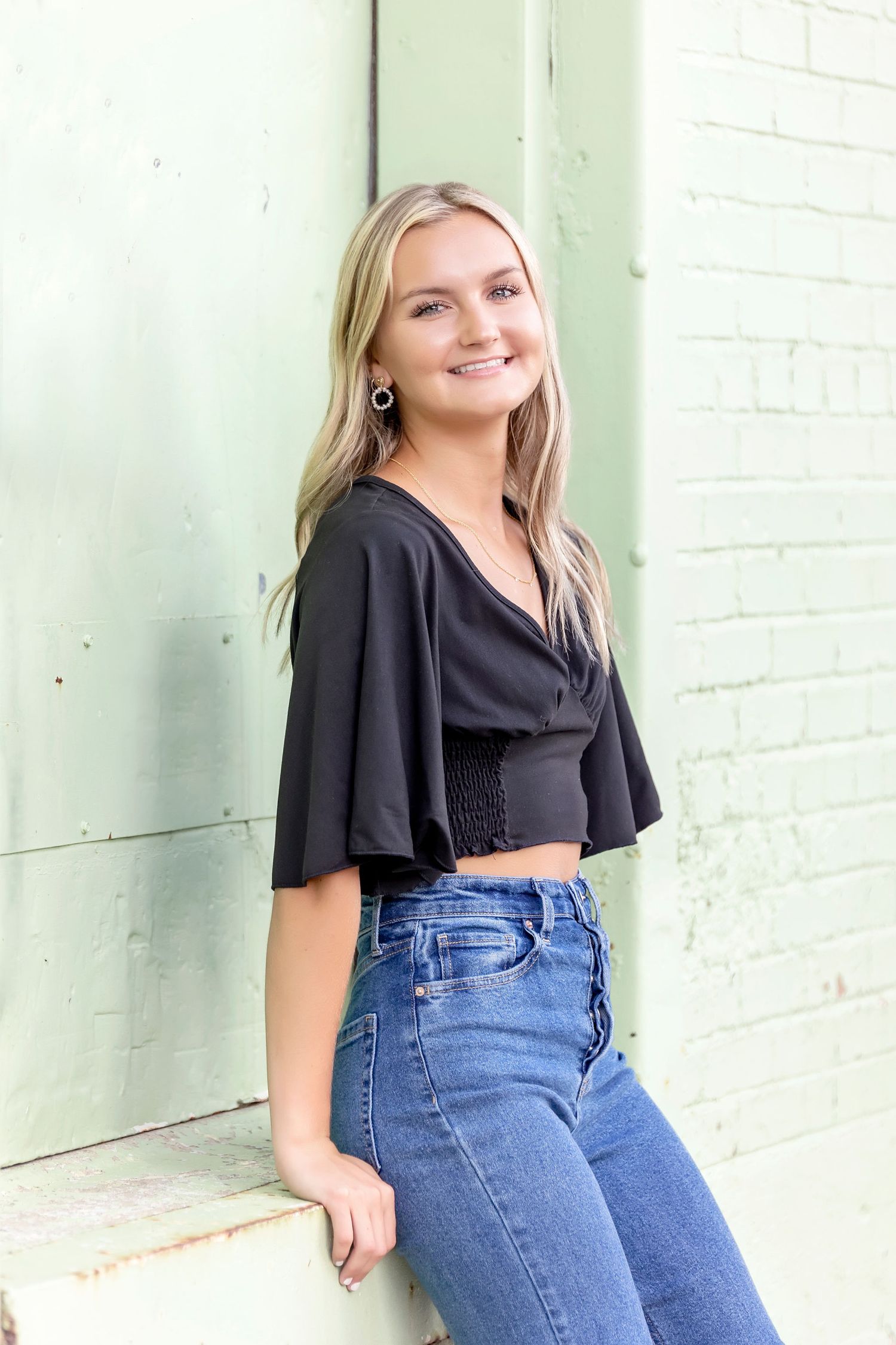 Casual senior portrait outdoors in Mason City against mint-green brick wall wearing jeans and black top